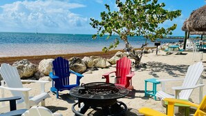 A beachside seating area with Adirondack chairs in various colors, a fire pit, and a thatched-roof structure in the background. - Refined. Safe. Clean. Waterfront. Golf cart , courtesy to our guests (Key Largo)