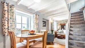 A dining area with a wooden table and chairs, a bowl of fruit, and a view of the outdoors through a window. - Cosy  barn close to Praa Sands (Helston)