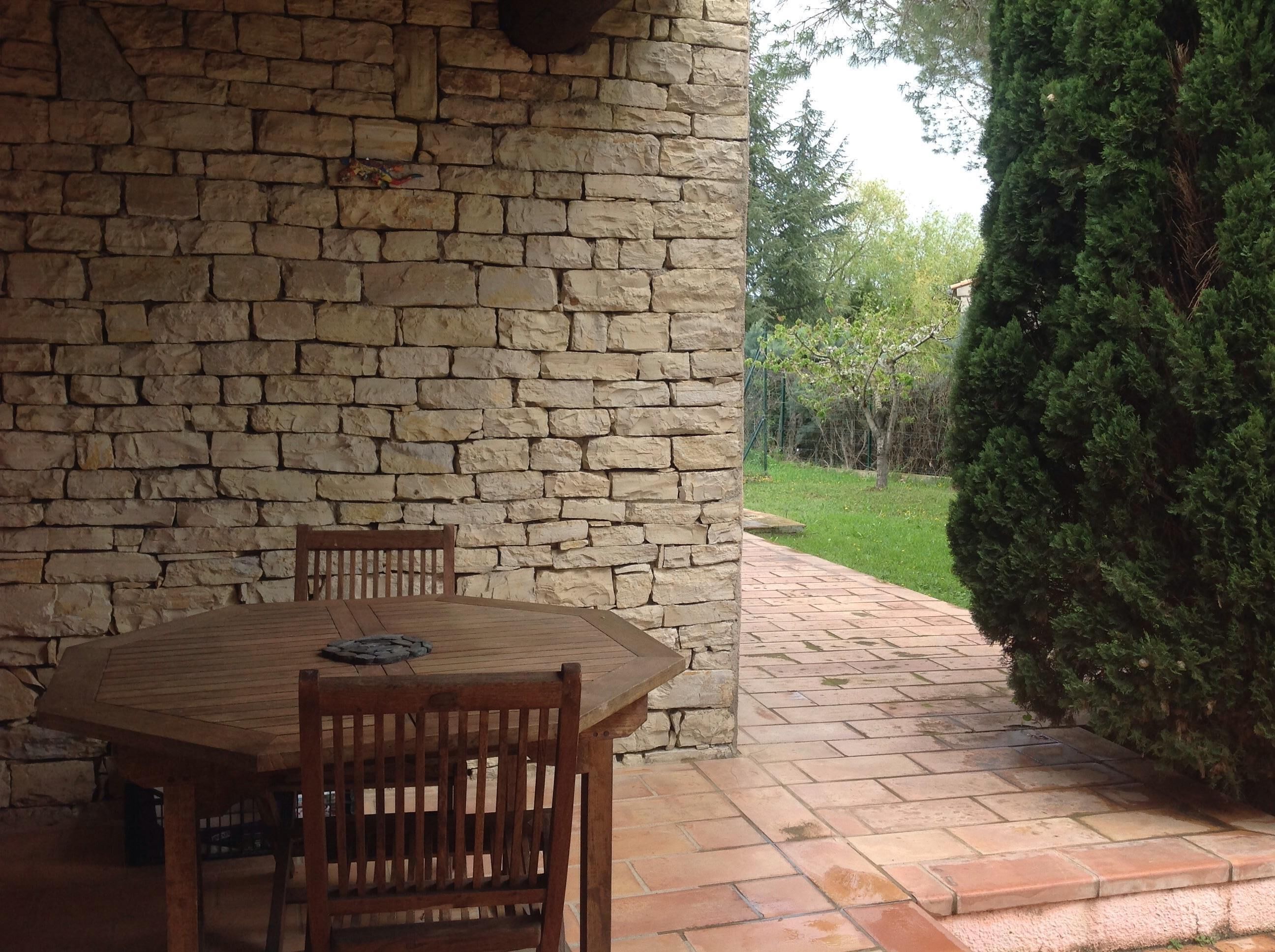 A stone wall with a wooden table and chairs, a stone bench, and a pathway leading to a green area with trees.
