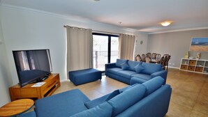 Ein Wohnzimmer mit Ecksofa, einem Fernseher auf einem Holzboden, einem Essbereich mit Stühlen und Blick durch eine Glastür.