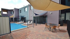 Ein Außenpoolbereich mit Liegestühlen und einem Tisch, umgeben von einer Pflasterterrasse und einem Gebäude mit großen Fenstern sowie einer überdachten Sitzgelegenheit.