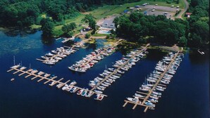An aerial view of a marina with numerous boats docked at wooden piers, surrounded by lush greenery and a small building with a swimming pool.