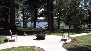 A park with a fire pit, benches, and a view of a lake with boats.