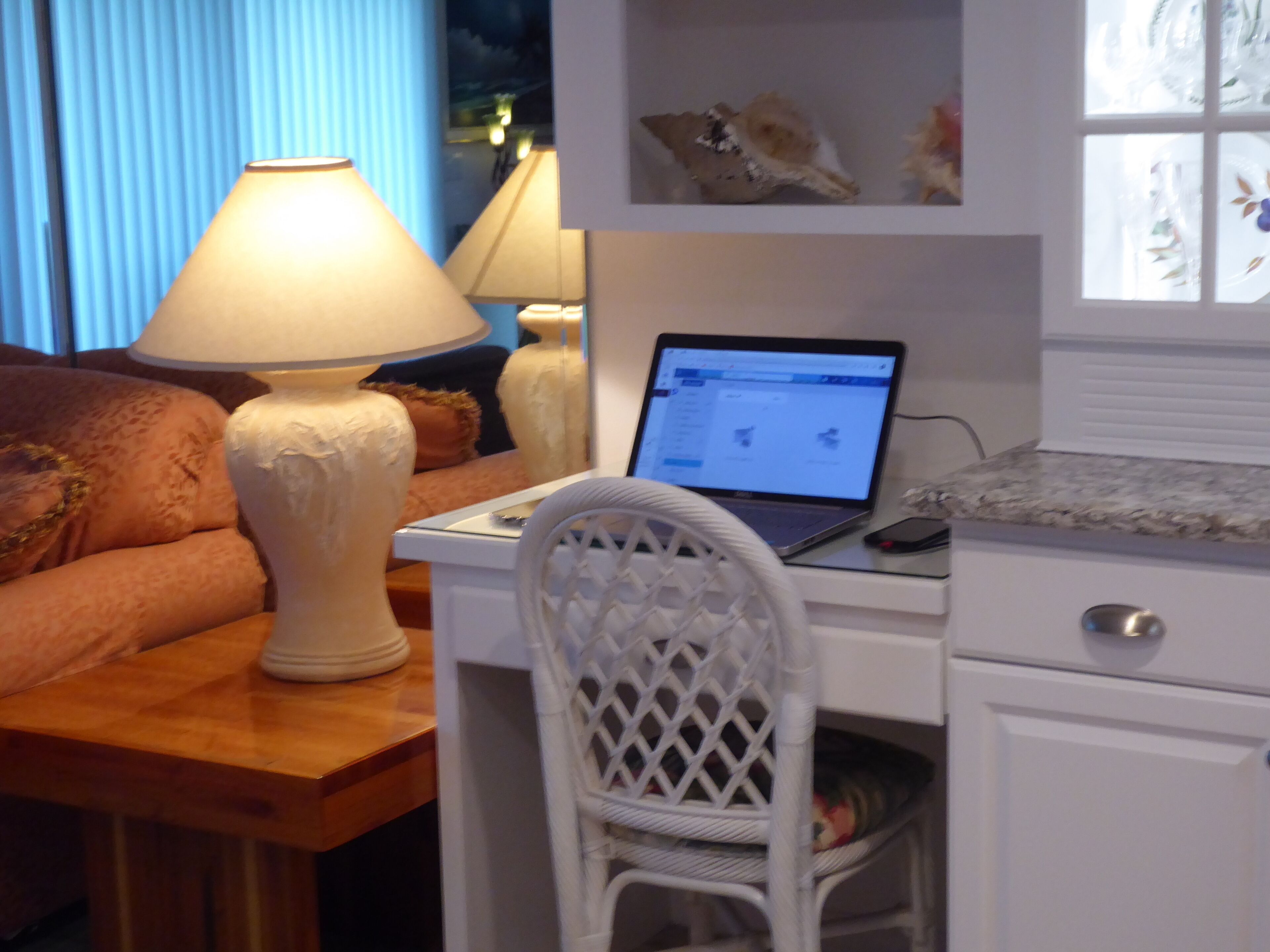 Een kamer met een houten bureau, een laptop, een lamp, een rieten stoel en een bed met een blauw gordijn.
