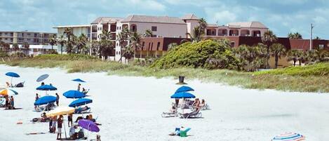 Una spiaggia con numerosi ombrelloni e lettini, un edificio con tetto rosso e palme sullo sfondo.