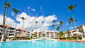 A swimming pool with lounge chairs and palm trees, surrounded by hotel buildings with red roofs. - Beautiful 2 bedroom 2 bath in a beachfront community (Punta Cana)