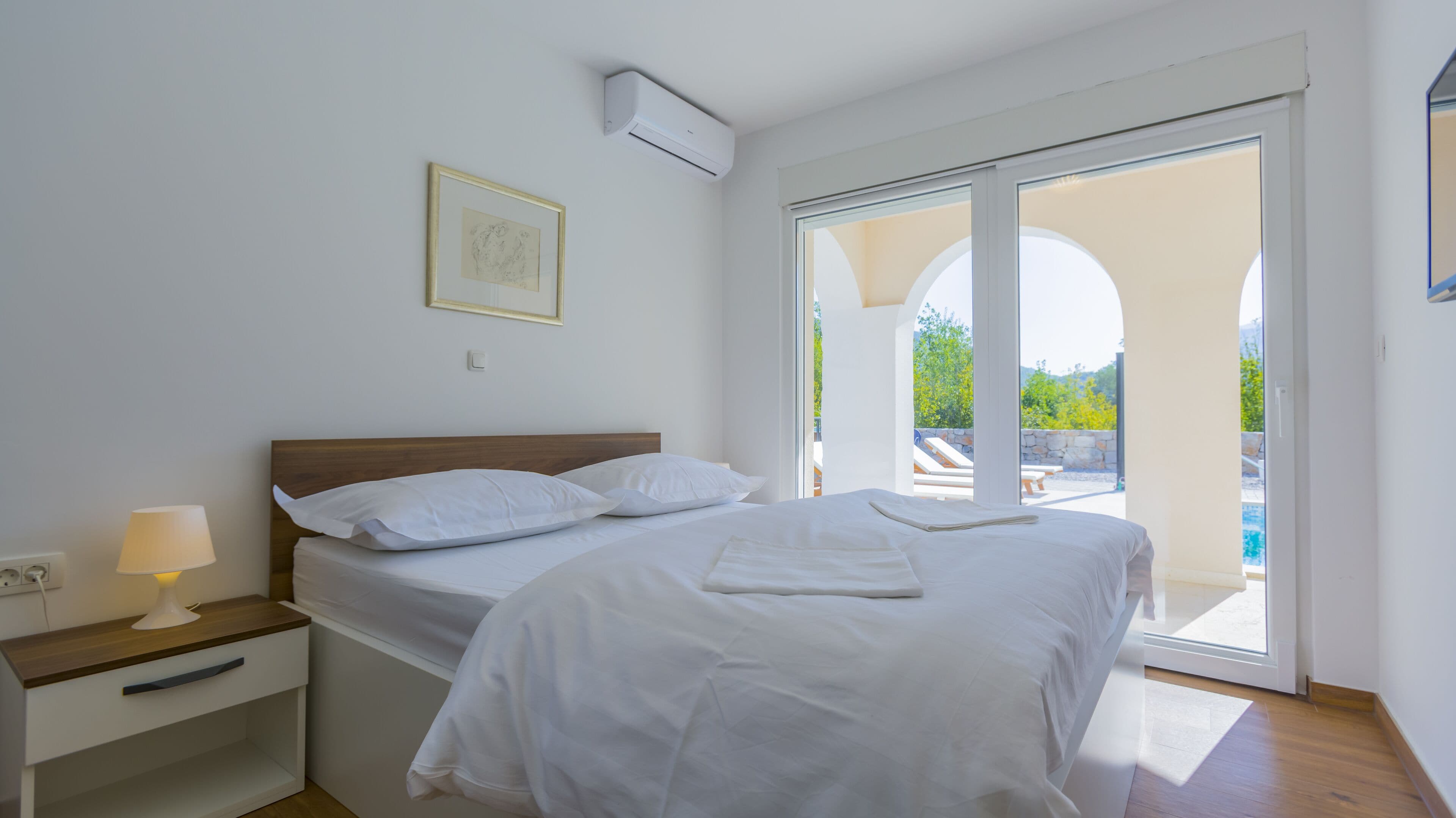 A bedroom with a bed, a bedside table with a lamp, a framed picture on the wall, and a sliding glass door leading to an outdoor area with a pool.