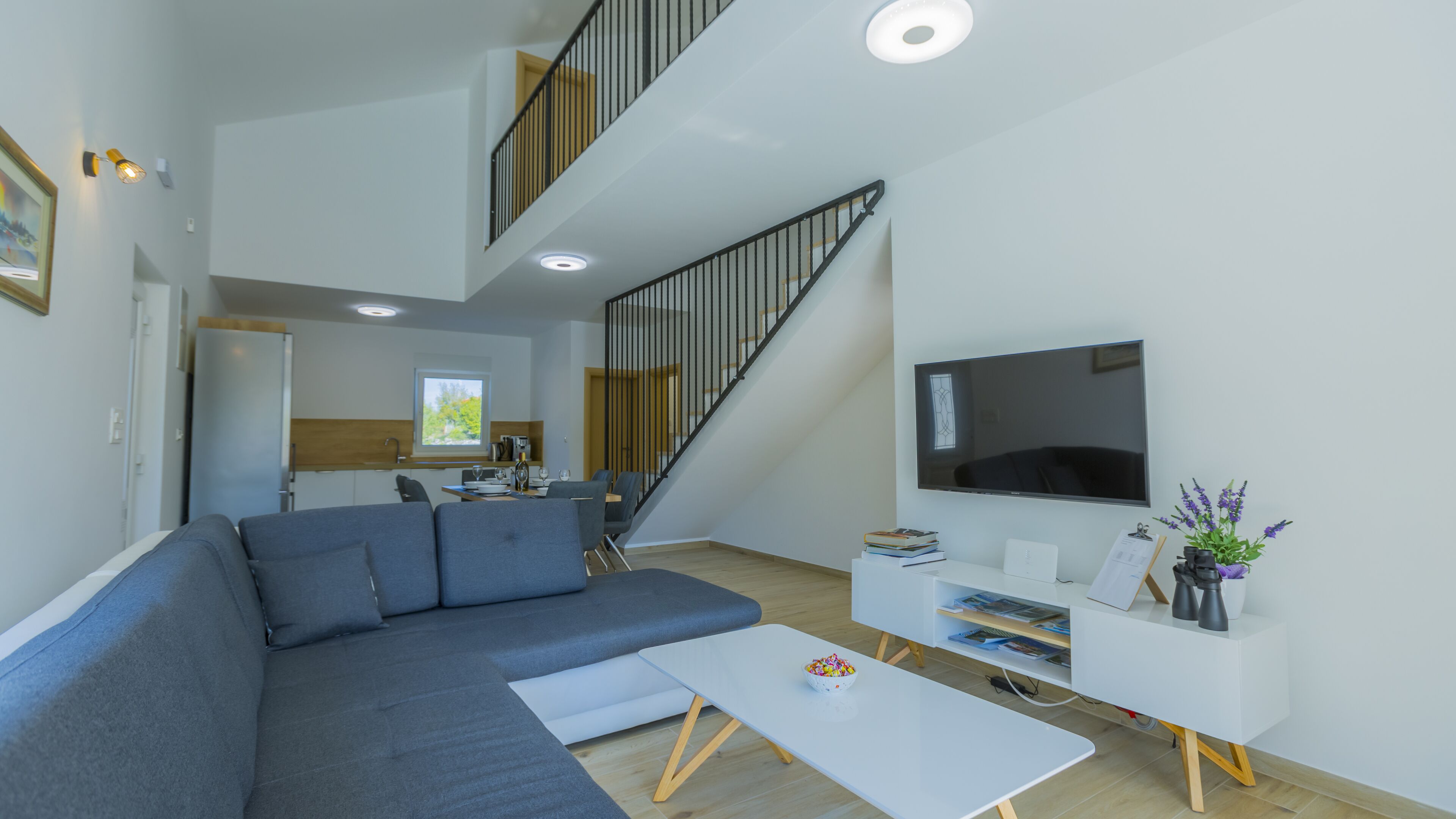 Un salon moderne avec un canapé d’angle gris, une table basse blanche et un téléviseur à écran plat posé sur un meuble blanc. Un escalier avec une rampe noire mène à l’étage supérieur.