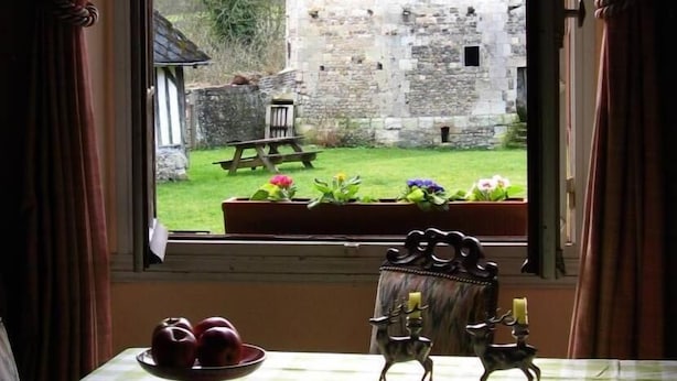 A view through a window of a stone building with a thatched roof, a wooden bench, and a flower bed.