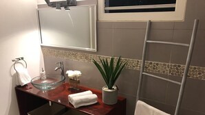 Un baño con un lavamanos de vidrio, un espejo, un perchero para toallas, una planta en maceta y una toalla en un perchero.