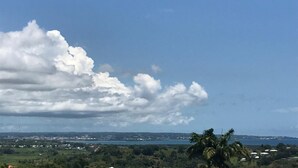 Un paisaje con cielo despejado, algunas nubes dispersas y una vista distante de la costa con edificios y vegetación.
