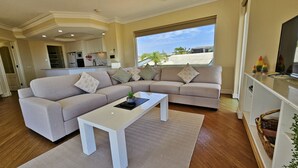 Un salon moderne avec un canapé d’angle beige, une table basse blanche et une grande fenêtre donnant sur le quartier.