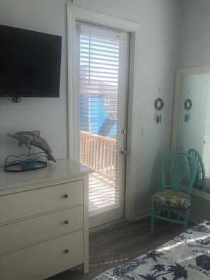 Una habitación con un aparador blanco, una silla verde azulada, un televisor montado en la pared y una puerta con persianas.
