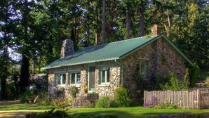 A stone cottage with a green roof, surrounded by a stone wall and flowering plants.