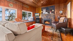 A cozy cabin interior with a stone fireplace, wooden walls, a large window, and a comfortable seating area.