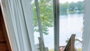 Una mesa pequeña con mantel floral, una copa de vino y un libro abierto junto a una ventana con vista a un lago.