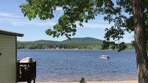 A lakeside view with a boat on the water, a small dock, and a building in the background. - Beautiful Views @ 4-Season Lake House, Private Sandy Beach (Fryeburg)