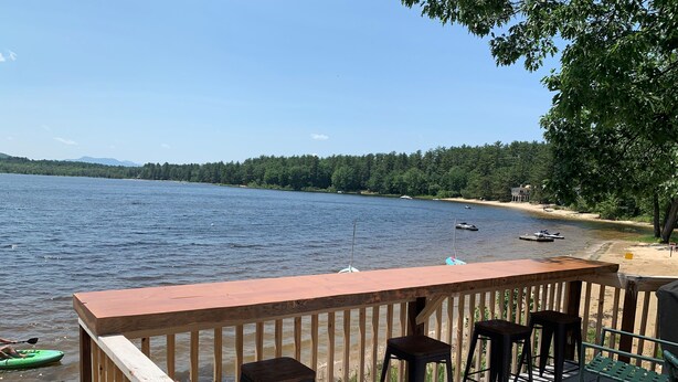 A wooden deck with a railing overlooking a lake, featuring a bar area with stools and a view of boats on the water. - Beautiful Views @ 4-Season Lake House, Private Sandy Beach (Fryeburg)