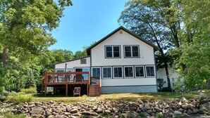 A two-story house with a deck, surrounded by trees and a stone wall. - Beautiful Views @ 4-Season Lake House, Private Sandy Beach (Fryeburg)