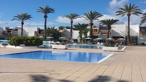 A swimming pool with lounge chairs and umbrellas, surrounded by palm trees and modern buildings. - Spring Apartment  (Costa del Silencio)