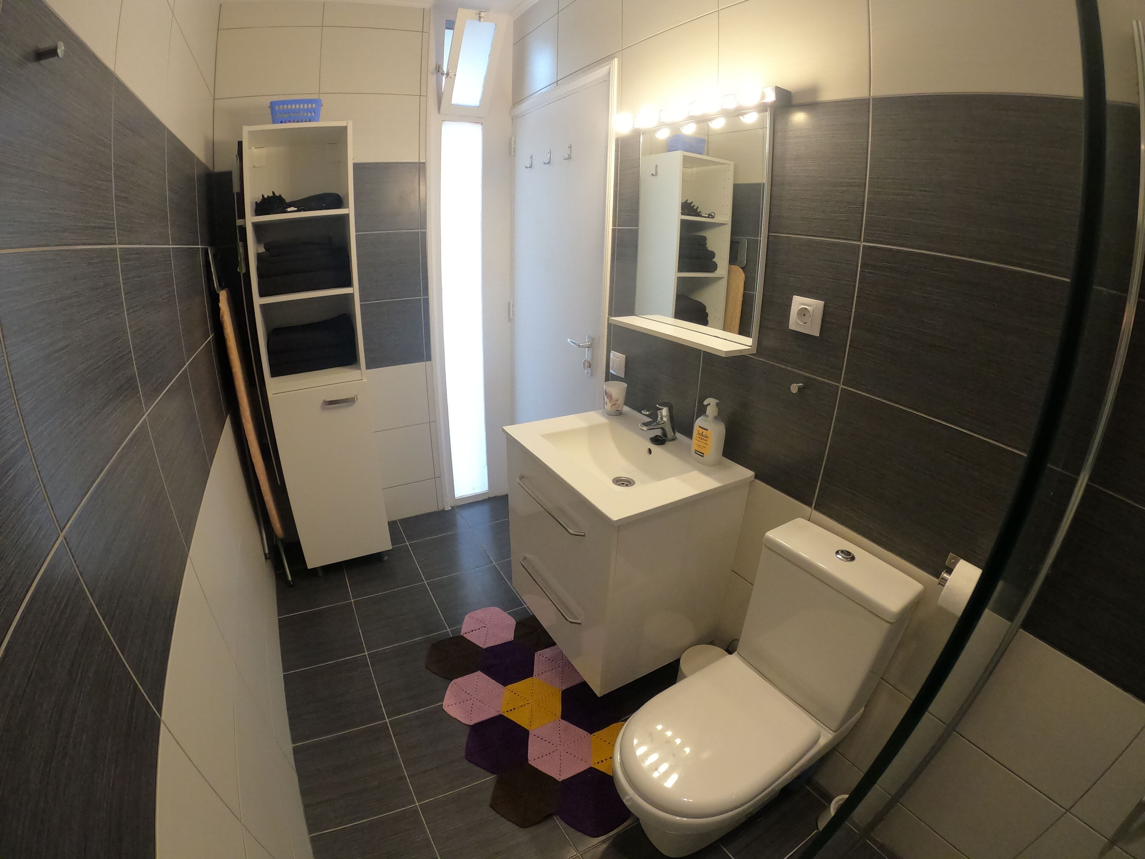 A modern bathroom with a white sink, mirror, and toilet. There is a storage cabinet with shoes and a small shelf. The floor has a geometric rug.