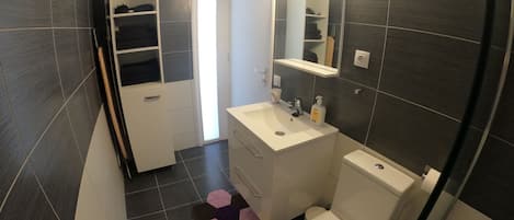 A modern bathroom with a white sink, mirror, and toilet. There is a storage cabinet with shoes and a small shelf. The floor has a geometric rug.