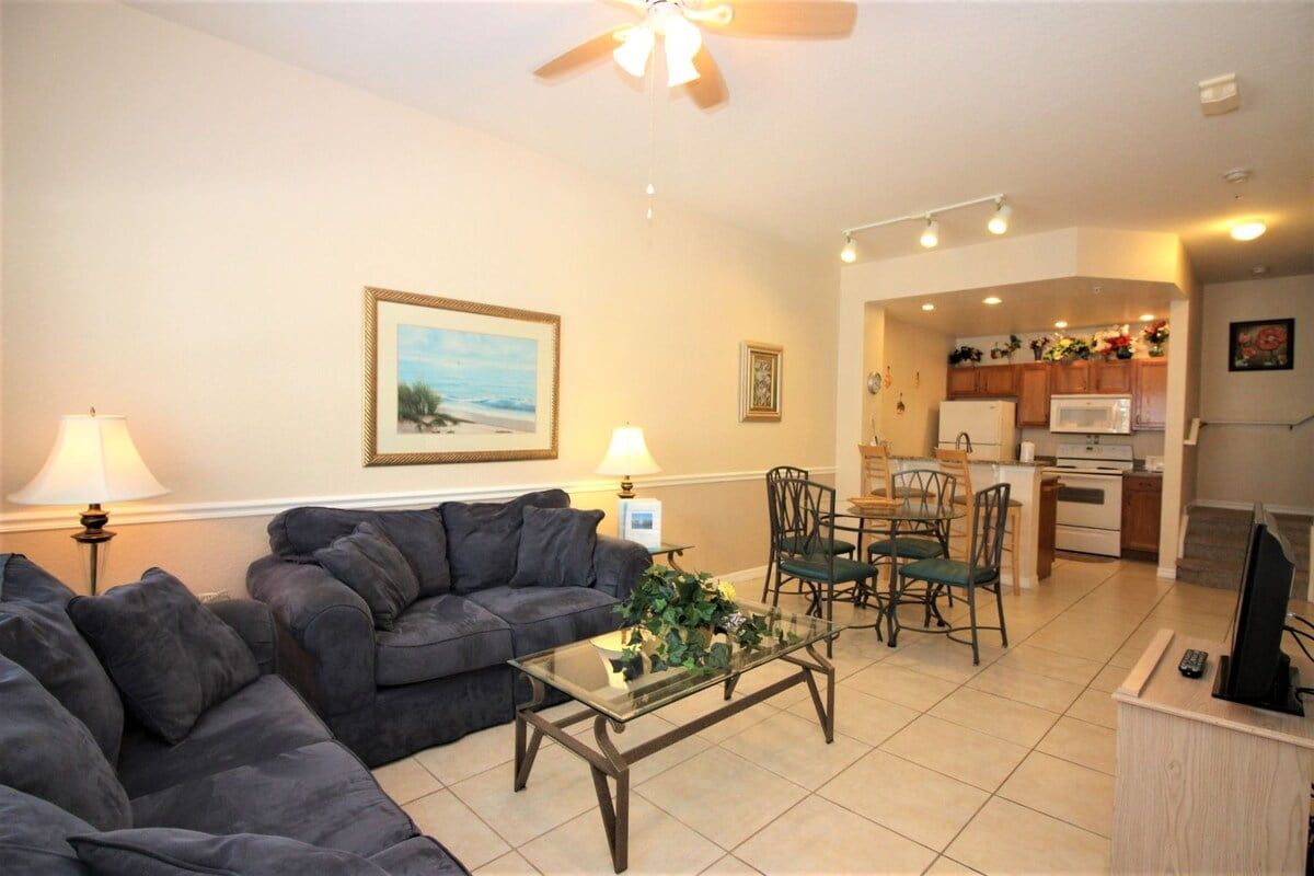 A living room with a sofa, coffee table, and a TV stand. There is a dining area with chairs and a table. The room has a ceiling fan and recessed lighting.