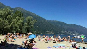 A crowded beach with people sunbathing and swimming, surrounded by mountains and clear blue water. - "FARFALLA" HOLIDAY HOUSE with PANORAMIC VIEW on LAKE MAGGIORE (Cannobio)