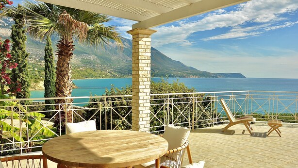 Une terrasse avec une table et des chaises en bois, une balustrade blanche et une vue sur la mer et les montagnes.