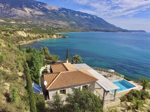 A coastal property with a stone house, a swimming pool, and a view of the sea and mountains.