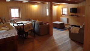 A cozy wooden cabin interior with a dining area, a sofa, a television mounted on the wall, and a window providing natural light. - MASET POZZATTI MOUNTAIN CHALET (Baselga)