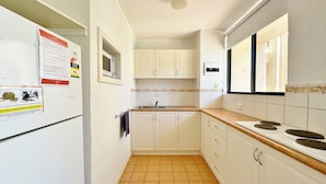 Une cuisine étroite avec des meubles blancs, un four à micro-ondes et une cuisinière. Le sol est carrelé et il y a une fenêtre avec un rideau.