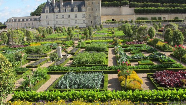 Un castillo histórico con un jardín formal que presenta parterres geométricos y una variedad de plantas con flores.