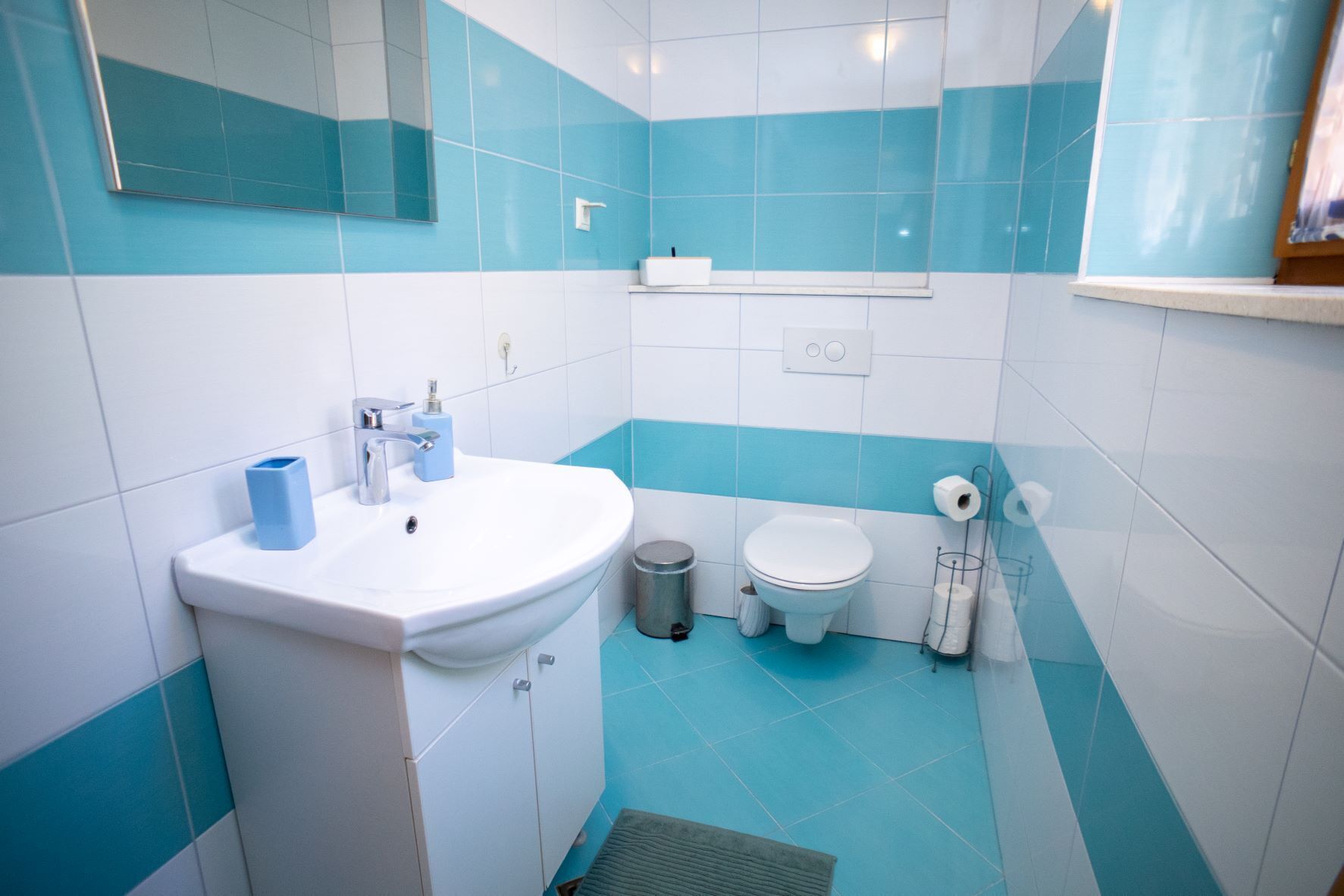A bathroom with a white sink, blue and white tiles, a toilet, and a trash can.