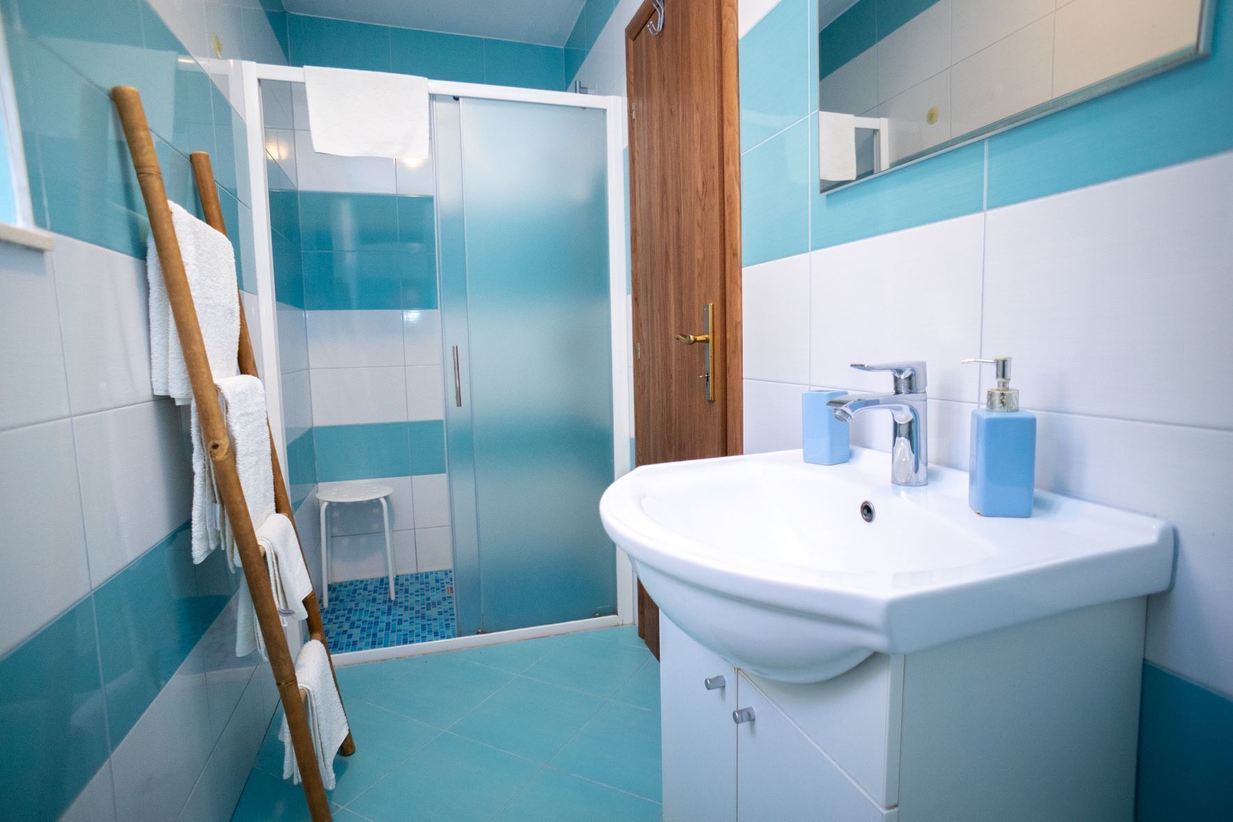 A bathroom with a white sink, blue tiles, a wooden door, and a glass shower enclosure.