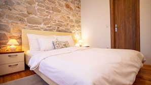 Un dormitorio con pared de piedra, puerta de madera, cama con ropa de cama blanca, dos lámparas de noche y una mesita de noche con cajones.