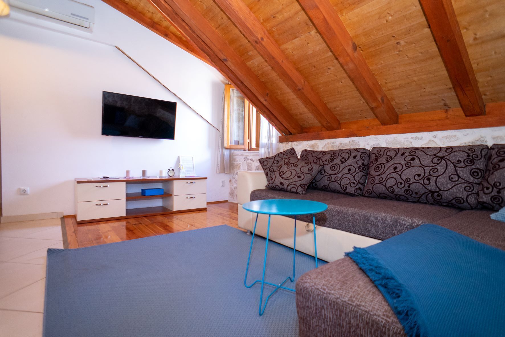 A cozy loft-style room with a wooden ceiling, a sofa with patterned cushions, a small blue table, and a TV mounted on the wall.