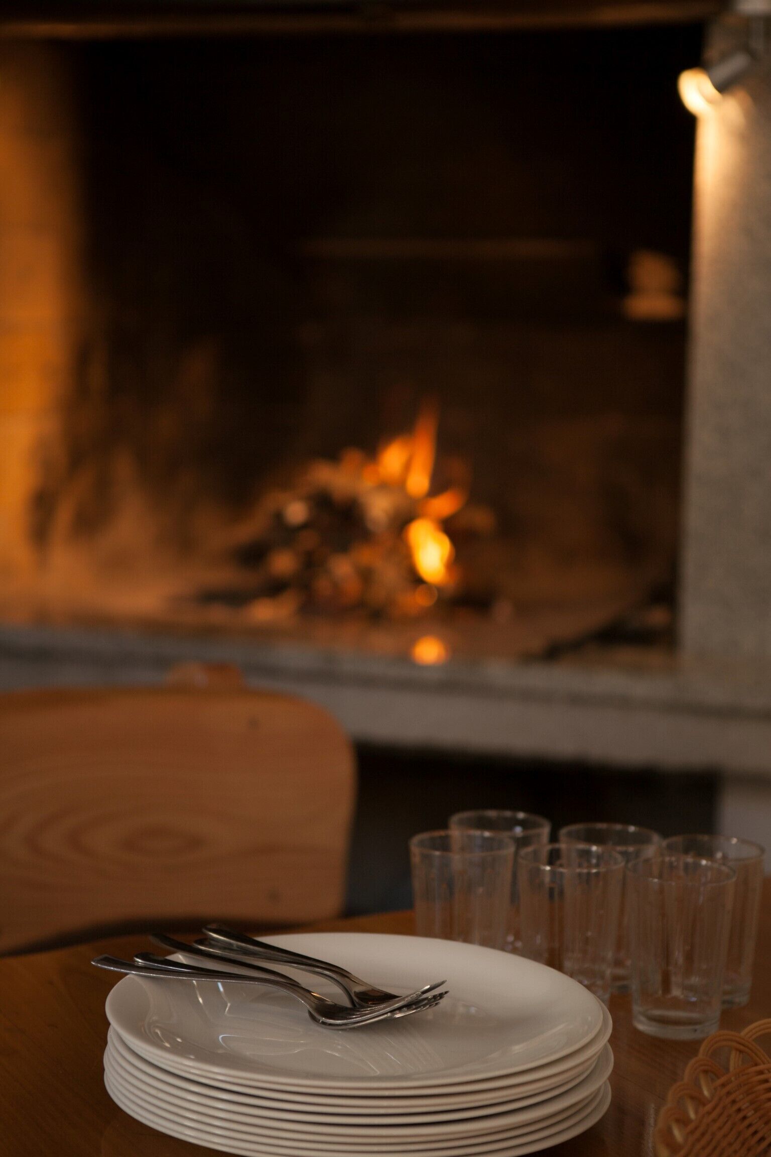 Una mesa de comedor con platos apilados y cubiertos, una chimenea con fuego a la vista, y varios vasos.