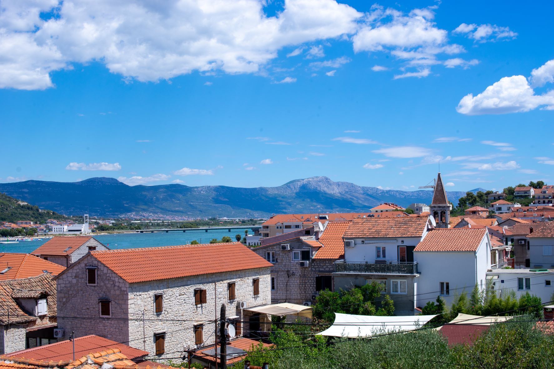 A coastal town with stone buildings and red-tiled roofs, a clear blue sky, and mountains in the background.