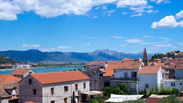 A coastal town with stone buildings and red-tiled roofs, a clear blue sky, and mountains in the background.