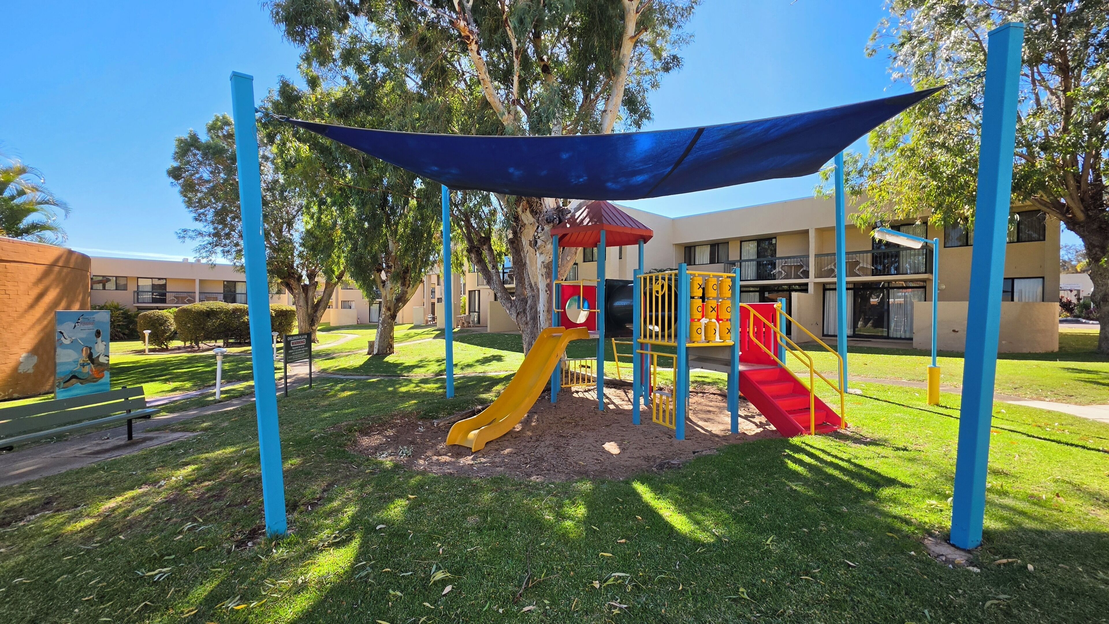 A playground with slides and climbing structures, surrounded by trees and a building in the background.