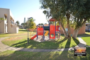Playground area with a slide and climbing structure, surrounded by a grassy lawn and shaded by trees.