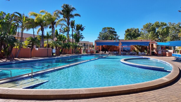 A swimming pool with a curved design, surrounded by palm trees and a covered seating area. - Riverview Holiday Waterfront Apt 25 - 1st Floor - Kalbarri WA (Kalbarri)