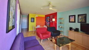 A living room with a purple sofa, a red and a black chair, a glass coffee table, a ceiling fan, and a TV on a wooden cabinet. - Riverview Holiday Waterfront Apt 25 - 1st Floor - Kalbarri WA (Kalbarri)