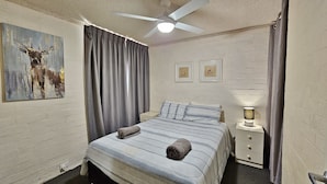 Een slaapkamer met een bed, een plafondventilator, een nachtkastje met een lamp, en twee ingelijste foto's aan de muur.