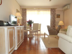 Uma sala de estar com televisão, mesa de jantar com cadeiras, sofá e uma cadeira de vime.