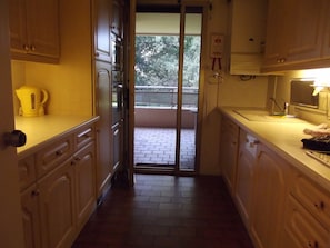 Cozinha estreita com armários de madeira, chaleira no balcão e vista para uma varanda com árvores.