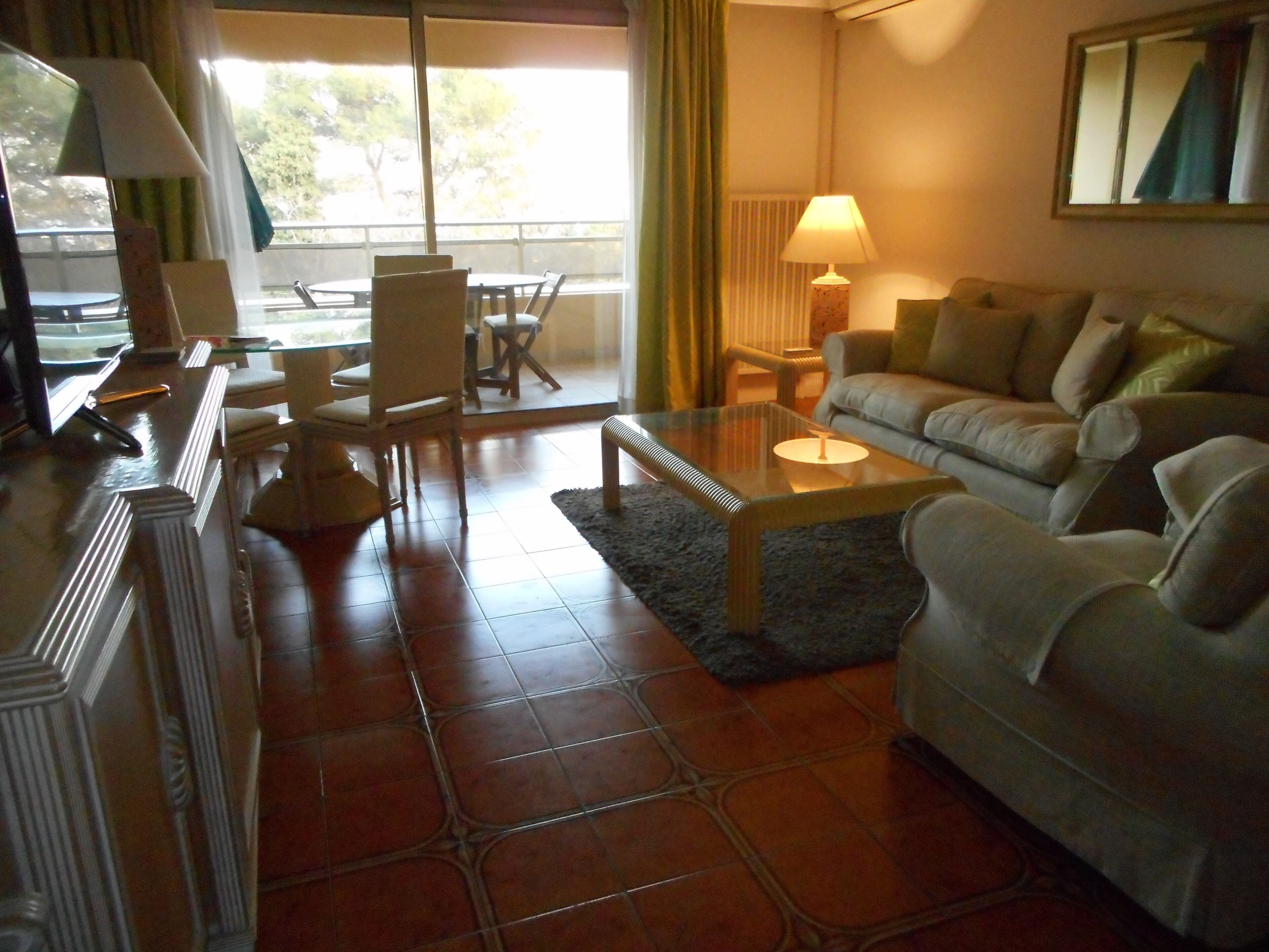 Un salon comprenant un canapé, des fauteuils, une table basse et un téléviseur. Il y a une fenêtre avec des rideaux, une lampe et une vue sur l’extérieur.