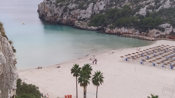 A beach with a rocky cliff, clear turquoise water, and a sandy area with umbrellas and beach chairs. - Casa Llebeig - Apartament with panoramic Sea view of Cala en Porter - Menorca (Cala en Porter)