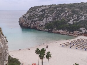 Une plage bordée d’une falaise rocheuse, avec une eau turquoise et limpide, et une zone de sable où l’on trouve des parasols et des chaises de plage.